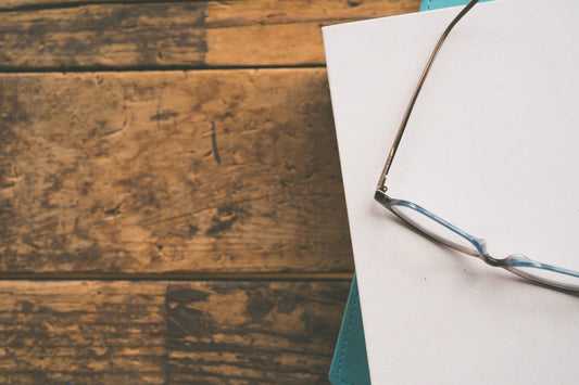 Minimalist composition of eyeglasses on white paper atop a wooden surface, perfect for lifestyle and office-themed visuals.