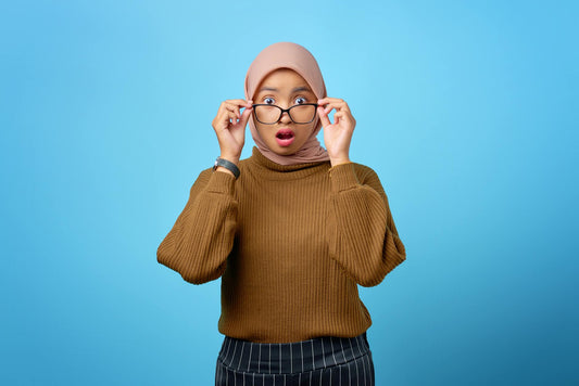 Asian woman in hijab wearing glasses with a surprised expression against a blue background.