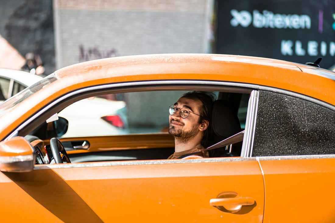 A cheerful young man with a beard and glasses smiles while driving an orange car outdoors.