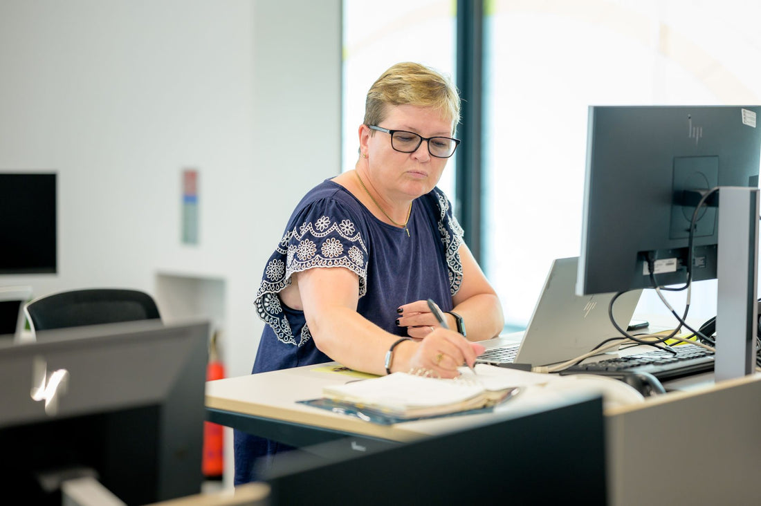 Professional woman wearing eyeglasses working at an office desk with a laptop and notebook.