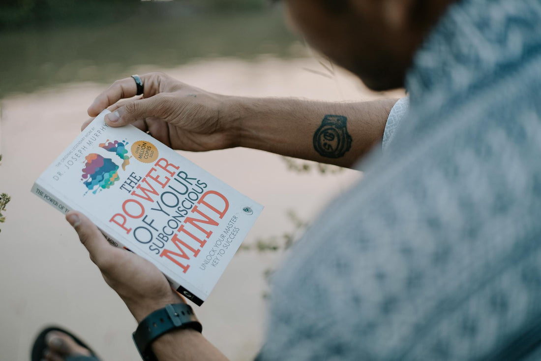 A man reads 'The Power of Your Subconscious Mind' by a riverside, showcasing relaxation and reflection.