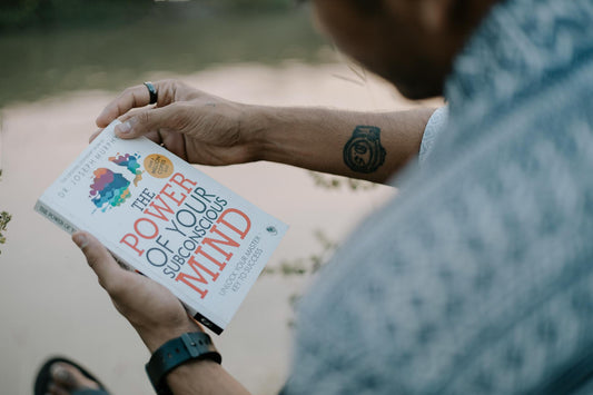A man reads 'The Power of Your Subconscious Mind' by a riverside, showcasing relaxation and reflection.