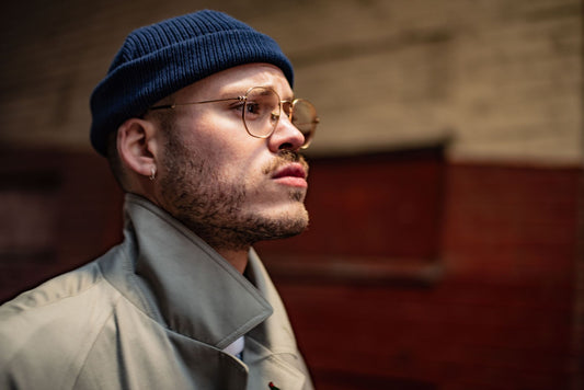 Stylish man with beard and glasses wearing a beanie and trench coat in an urban setting.