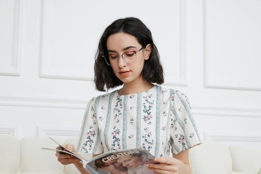 A young woman with glasses reads a magazine in a bright, minimalistic room, showcasing relaxed lifestyle and fashion.