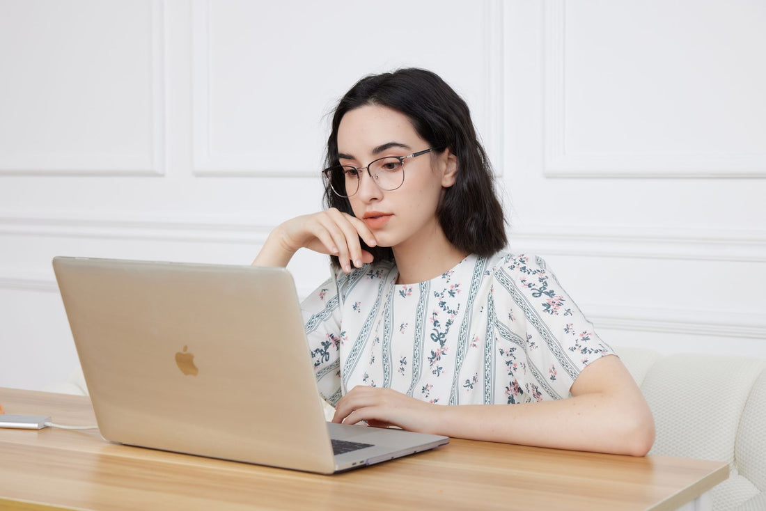 Woman in floral blouse wearing glasses, focused on laptop work at home.