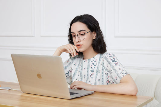 Woman in floral blouse wearing glasses, focused on laptop work at home.