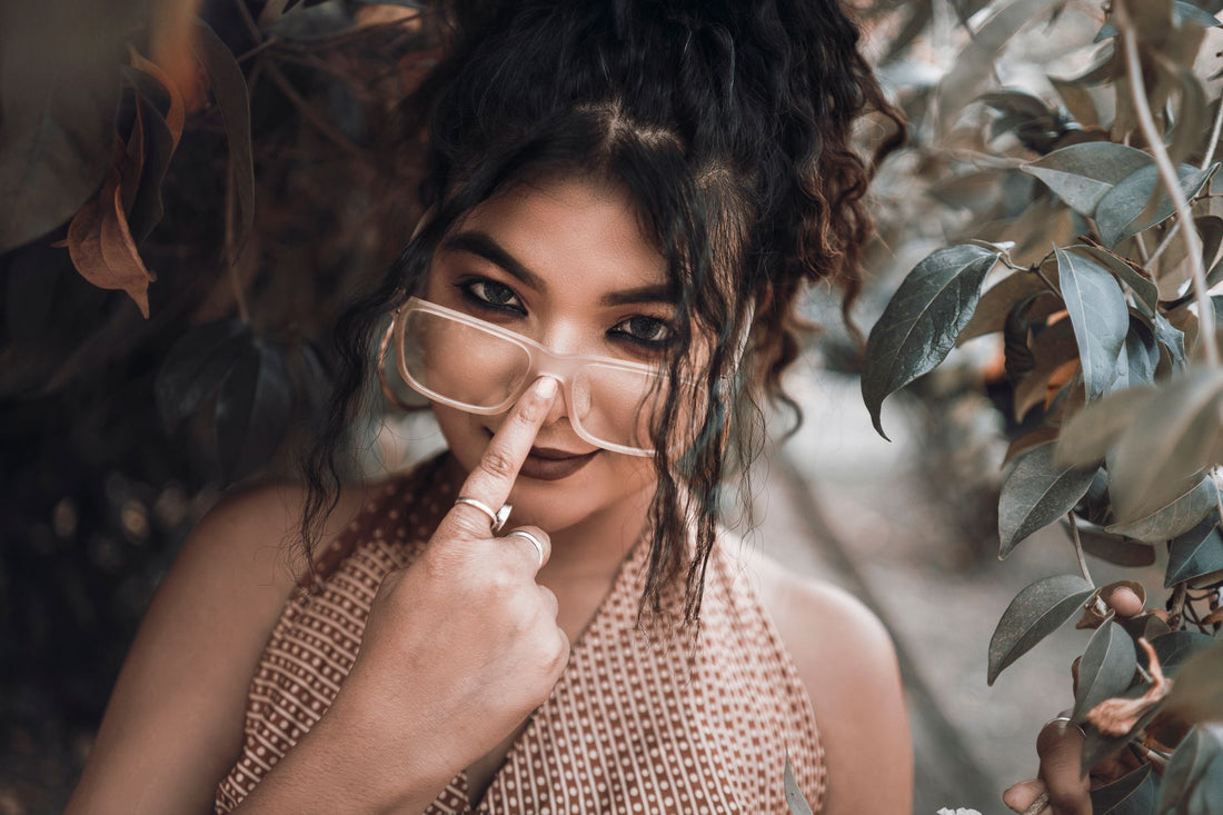 South Asian woman playfully adjusting glasses amidst leafy surroundings.