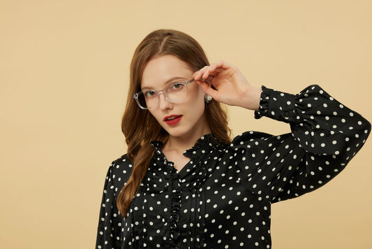 A fashionable woman wearing eyeglasses and a polka dot blouse poses against a beige background.