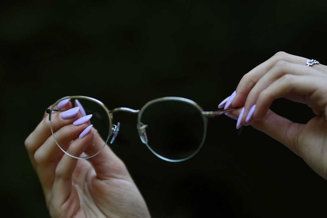 Close-up of hands with manicured nails holding eyeglasses against a dark background.