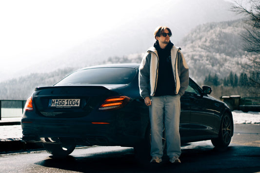 A man stands next to a sleek car in a scenic snowy mountainous area.