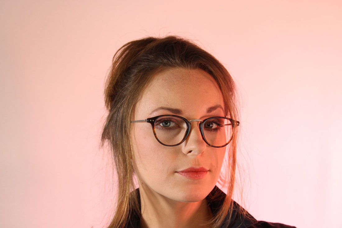 Close-up portrait of a woman wearing glasses, set against a soft pink background.