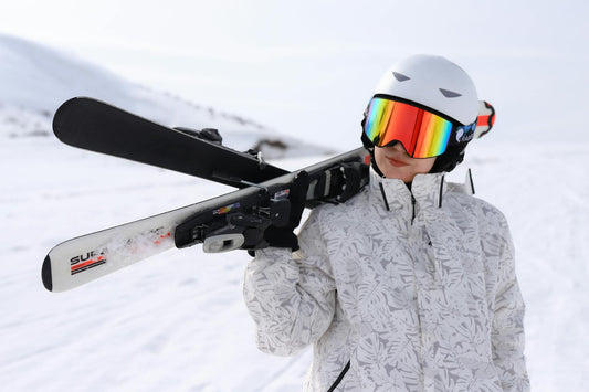 Person in ski gear holding skis over shoulder in snowy mountain setting, ready to hit the slopes.
