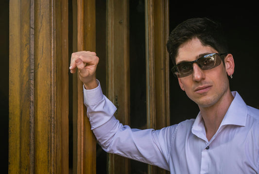 Confident man in sunglasses leaning on wooden door frame, wearing a white shirt.