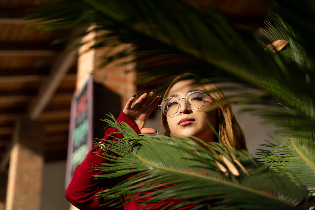 A stylish woman in glasses poses behind a lush fern indoors, illuminated by warm sunlight.