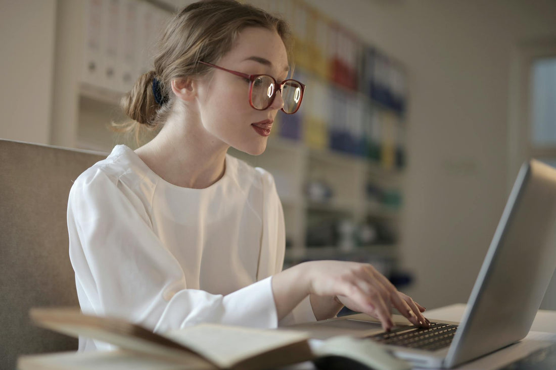 Focused young woman wearing glasses, typing on a laptop indoors, emphasizing modern work life.