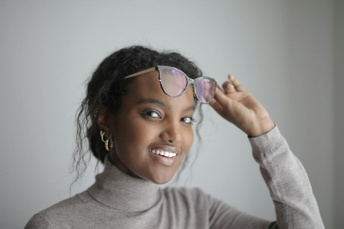 Portrait of a smiling woman in a turtleneck sweater adjusting her glasses indoors.