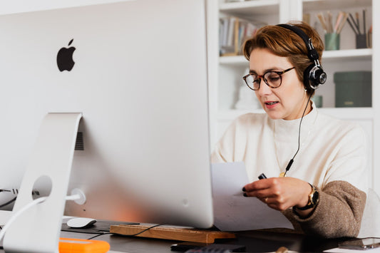 Content serious female in stylish sweater with headphones and eyeglasses having conversation via video call on computer and looking at papers while working in modern workspace