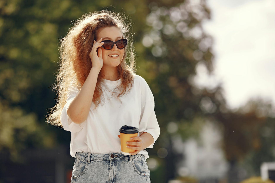 A woman with sunglasses and blonde hair enjoys a coffee in a sunny park.
