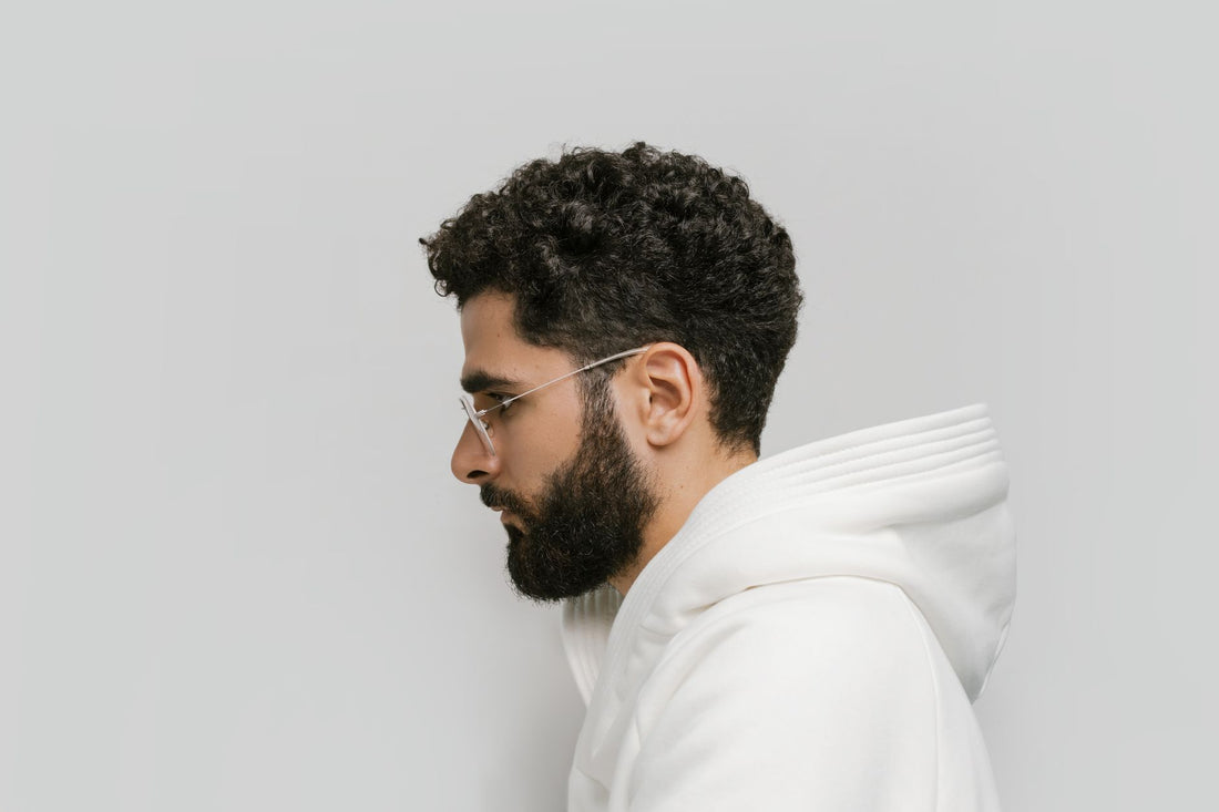 Side view of a bearded man with eyeglasses, wearing a white hoodie. Minimalist background.