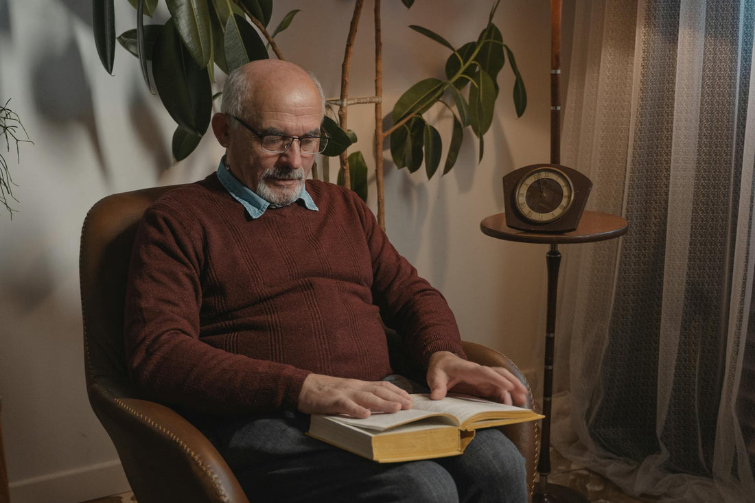 An elderly man reading a book in a cozy indoor setting, surrounded by plants and a vintage clock.