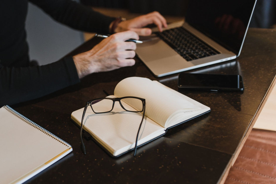 Professional setting with hands typing on laptop, notebook, and eyeglasses on table.