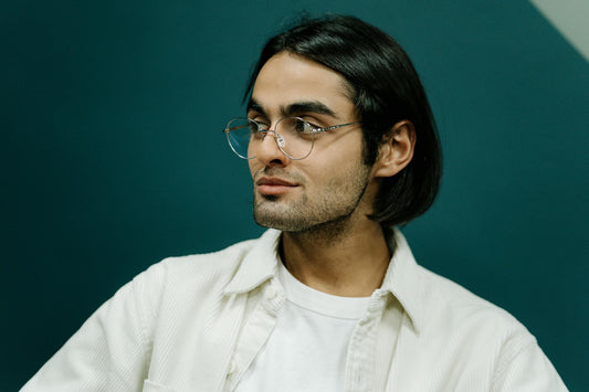 A thoughtful portrait of a stylish man wearing eyeglasses against a teal background.