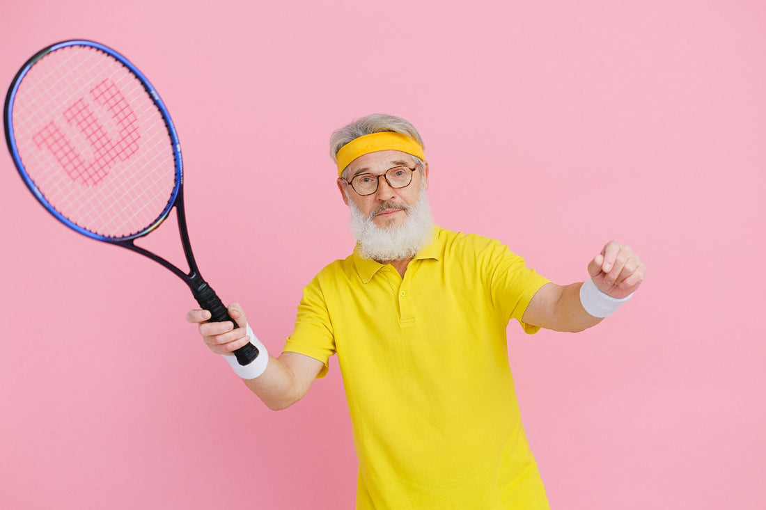 Elderly man in yellow activewear posing with a tennis racket against a pink background.
