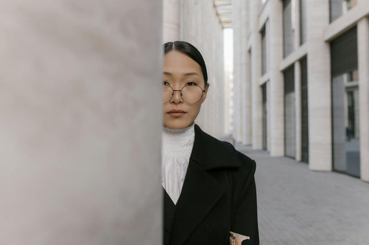 Elegant woman in black coat and eyeglasses stands in modern urban architecture.