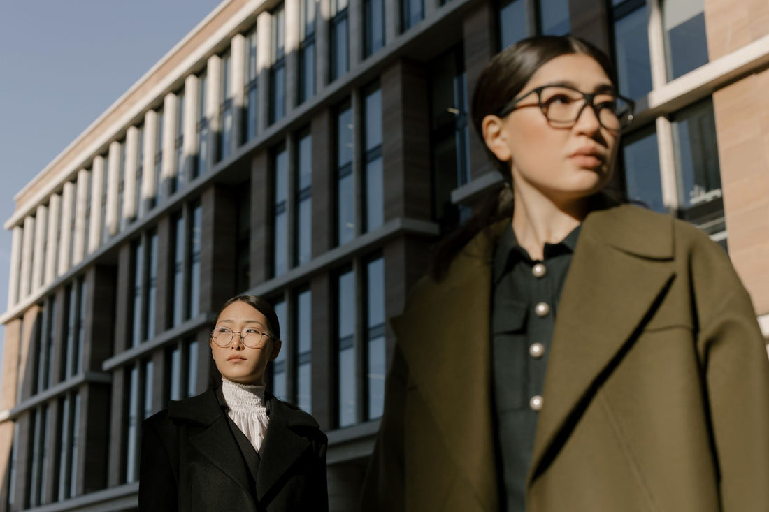 Two fashionable Asian women in stylish coats, wearing glasses, walking in an urban area.