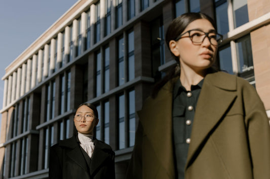 Two fashionable Asian women in stylish coats, wearing glasses, walking in an urban area.