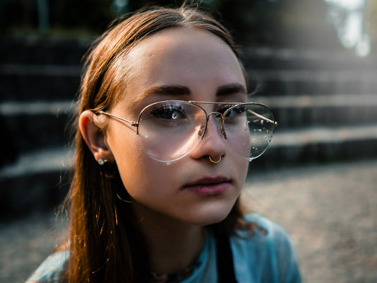 Close-up portrait of a young woman wearing glasses with water droplets, set outdoors in soft lighting.