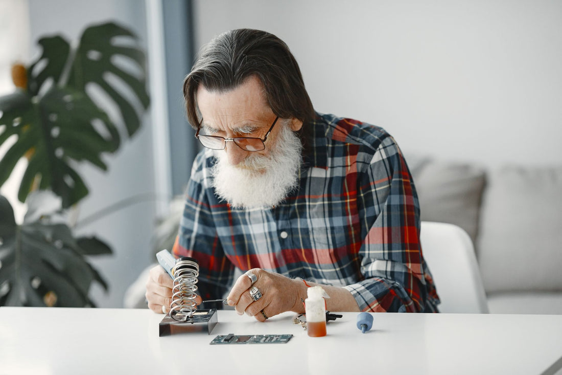 Elderly man with a beard working on electronics repair at a table indoors.