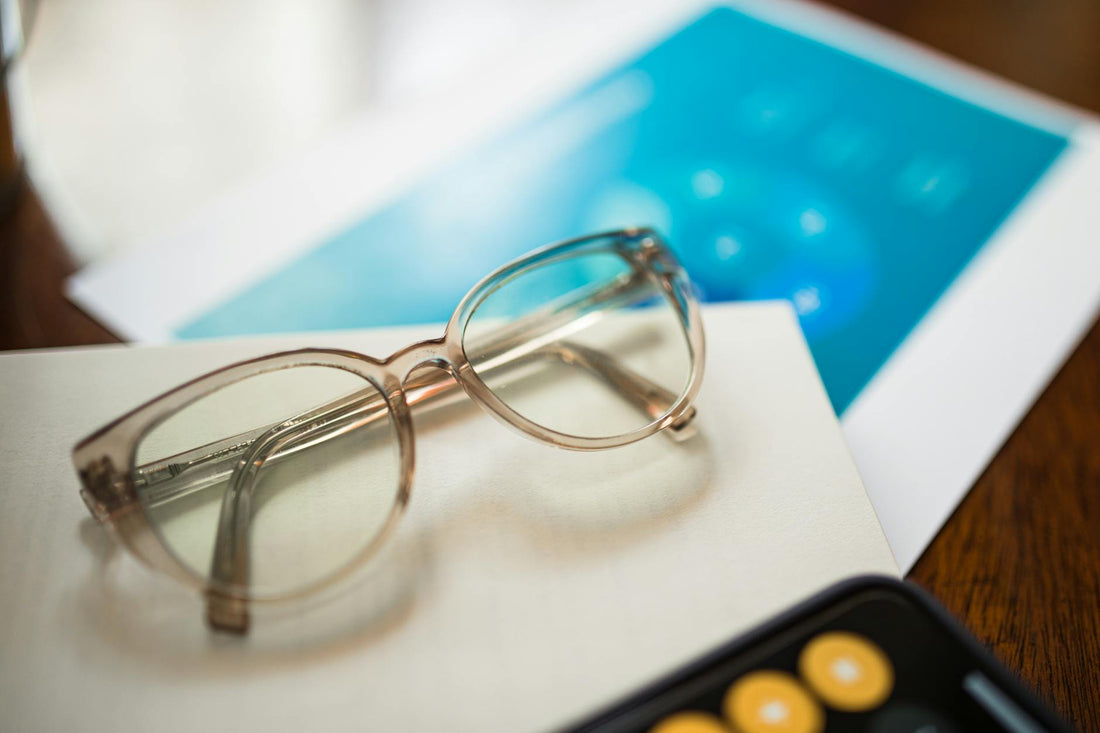 Close-up of eyeglasses on a desk with paper and blurred background, emphasizing minimalism.