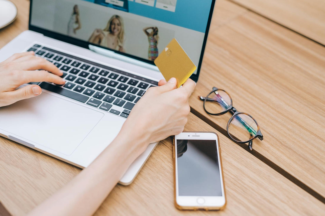 A person makes an online purchase using a credit card on a laptop, with a smartphone and glasses nearby.