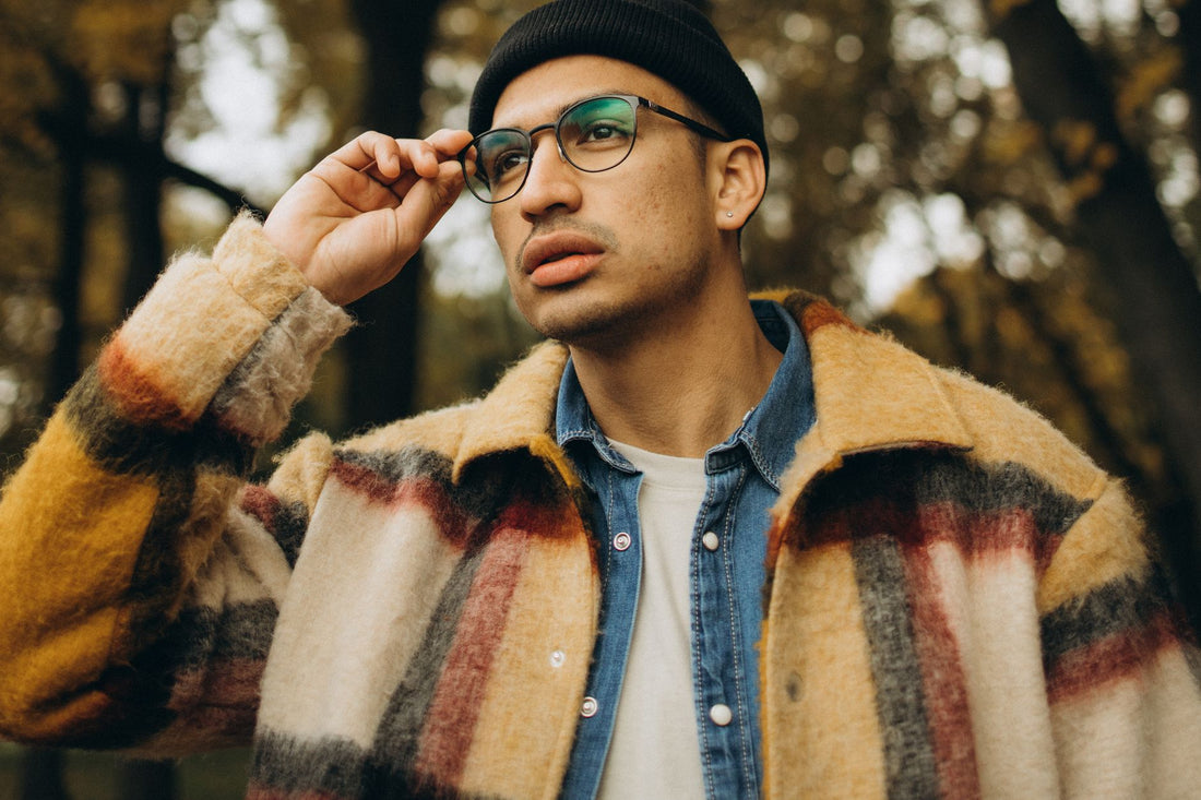 A fashionable man with eyewear and a checkered coat thoughtfully posing in a serene fall setting.